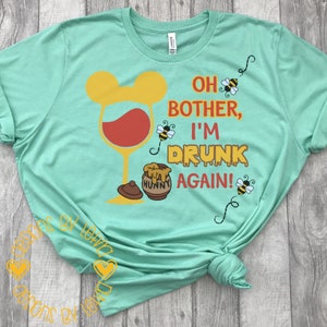 Könnte beinhalten: Ein türkisfarbenes T-Shirt mit dem Text "Oh bother, I'm drunk again!" Der Text ist in Rot und Gelb. Es gibt ein Glas Wein mit einem Mickey-Mouse-Ohren-Design und einen Honigtopf mit dem Wort "Hunny" darauf. Es gibt auch Bienen, die um das Design herumfliegen.