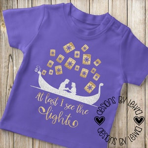 May include: Purple t-shirt with a gold glitter design of a couple in a boat with lanterns floating above them. The text "At last I see the light" is written below the boat. The text "designs by leiva" is written in the bottom right corner.