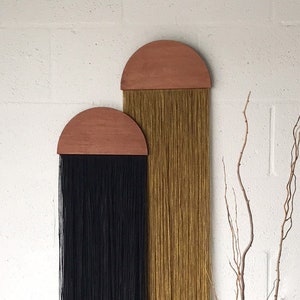 Könnte beinhalten: Zwei Wandbehänge mit halbrunden Holz-Oberteilen und langen Garnsträngen. Der linke Behang hat schwarzes Garn und der rechte Behang hat goldenes Garn. Eine schwarze Vase mit einem Trockenblumenarrangement steht auf einer weißen Oberfläche.