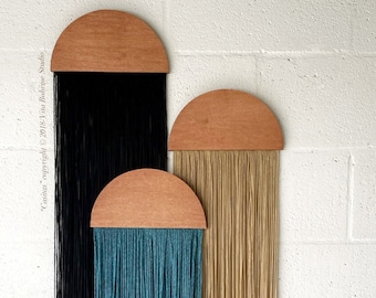 May include: Three wall hangings with wooden half-circle tops and long strands of yarn hanging down. The yarn is black, teal, and beige.