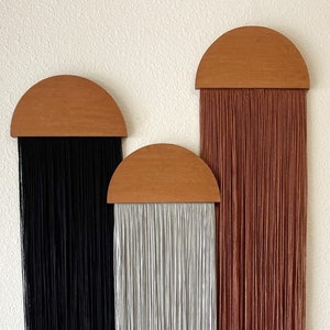 Könnte beinhalten: Drei Makramee-Wandbehänge mit halbrunden Holz-Oberteilen. Die Behänge sind in den Farben Schwarz, Grau und Rost.