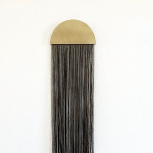 Könnte beinhalten: Ein schwarzer Holzhocker mit runder Oberseite und drei gedrehten Beinen. Die Beine haben ein dekoratives Design. Der Hocker steht vor einer weißen Wand mit einem grauen und goldenen Wandbehang.