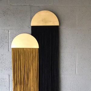 Könnte beinhalten: Zwei Wandbehänge mit goldenen Halbkreis-Oberteilen und langen Fransen. Die Fransen sind links schwarz und rechts senfgelb. Die Behänge befinden sich an einer grauen Backsteinwand.