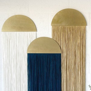 Könnte beinhalten: Drei Wandbehänge mit goldenen Halbkreis-Oberteilen und langen Garnsträngen. Die Stränge sind weiß, dunkelblau und beige.