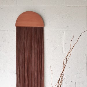 Könnte beinhalten: Ein brauner Makramee-Wandbehang mit einer halbrunden Holzspitze. Der Behang besteht aus vielen dünnen Garnsträngen und hängt an einer weißen Backsteinwand.