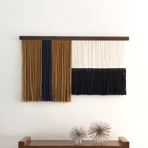 Könnte beinhalten: Ein Wandbehang aus Garn in den Farben Senfgelb, Marineblau und Weiß. Der Behang ist in vier Abschnitte unterteilt, wobei jeder Abschnitt eine andere Farbe aufweist. Der Behang ist an einer Holzleiste befestigt. Der Behang wird über einer Holzbank mit zwei silbernen Dekor-Kugeln präsentiert.