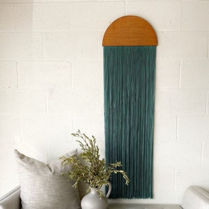 Könnte beinhalten: Eine weiße, gepolsterte Bank mit einem hellgrauen Kissen und einem Kissen. Eine Vase mit getrockneten Zweigen steht auf der Bank vor einem türkisfarbenen Wandbehang aus Holz.