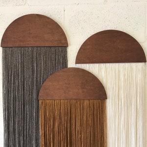 Könnte beinhalten: Drei halbkreisförmige Wandbehänge aus Holz mit langen Garnsträngen, die von unten hängen. Das Garn ist grau, braun und weiß.