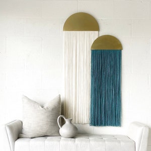 Könnte beinhalten: Eine weiße Bank mit einem hellgrauen Kissen und einer grauen Keramikvase. Zwei Wandbehänge mit goldenen Halbkreis-Oberteilen und Fransen hängen an der weißen Wand hinter der Bank.