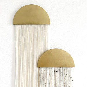 Könnte beinhalten: Zwei goldene Halbkreis-Wandbehänge mit weißen Garnsträngen, die nach unten hängen. Der obere Halbkreis hat schlichte weiße Garnstränge, während der untere Halbkreis weiße Garnstränge mit schwarzen Flecken hat.