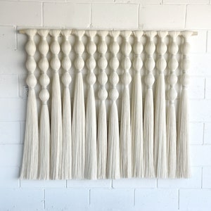 Könnte beinhalten: Ein weißer Makramee-Wandbehang mit einer Holzleiste oben. Der Behang besteht aus einer Reihe großer, geknoteter Schlaufen mit langen, fließenden Garnsträngen.