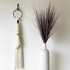 Könnte beinhalten: Eine weiße Keramikvase mit beigefarbenem Boden steht auf einem schwarzen Holzständer. Die Vase ist mit getrocknetem braunem Gras gefüllt.