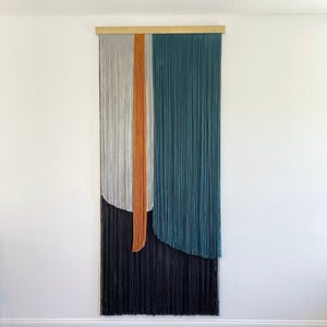 Könnte beinhalten: Ein Wandbehang aus Garn in den Farben Grau, Orange, Türkis und Schwarz. Das Garn ist zu einem geometrischen Muster mit geschwungenen Linien gewebt.