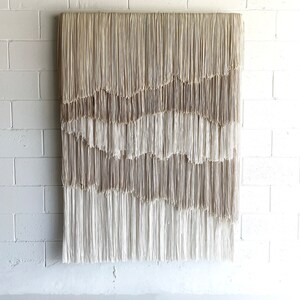 Op de afbeelding: Een groot wandkleed gemaakt van geweven beige en wit garen met een franje rand. Het garen is in een golvend patroon gerangschikt, waardoor een textuur en een moderne uitstraling ontstaat.