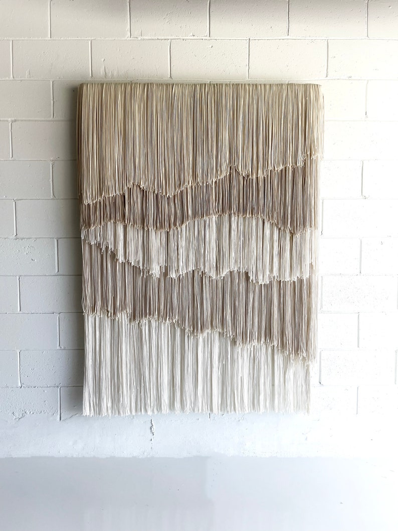 Op de afbeelding: Een groot wandkleed gemaakt van geweven beige en wit garen met een franje rand. Het garen is in een golvend patroon gerangschikt, waardoor een textuur en een moderne uitstraling ontstaat.