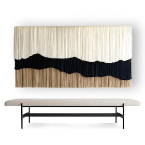 Könnte beinhalten: Ein großer, rechteckiger Wandbehang mit einem geschichteten Design. Der obere Teil ist cremefarben, gefolgt von einem schwarzen Band und einer beigefarbenen Franse am unteren Rand. Darunter eine lange Bank mit hellfarbenem Kissen und schwarzen Metallbeinen.