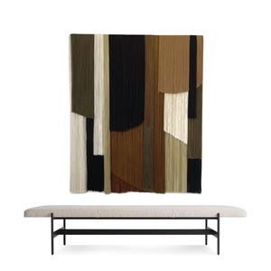 May include: A long, rectangular bench with a light gray fabric cushion and a black metal frame. The bench is in front of a wall hanging made of colorful yarn strands in shades of brown, green, and white.
