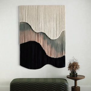 Könnte beinhalten: Ein großer, rechteckiger Wandbehang mit einem geschichteten Design. Der obere Bereich ist cremefarben, gefolgt von einem Bereich in gedämpftem Grün und Rosa und einem unteren Bereich in Schwarz. Eine grüne Bank und ein kleiner Holztisch sind im Vordergrund.