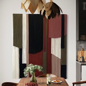 May include: A wooden dining table with a pizza, a glass of lemonade, a bowl of red berries, and a vase of white flowers. The table is set against a wall with a colorful macrame wall hanging and two wooden pendant lights.