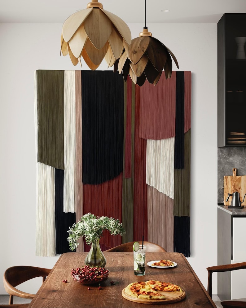May include: A wooden dining table with a pizza, a glass of lemonade, a bowl of red berries, and a vase of white flowers. The table is set against a wall with a colorful macrame wall hanging and two wooden pendant lights.