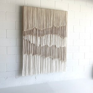 Op de afbeelding: Een grote wandhanger gemaakt van geweven beige en wit garen met franjes. De hanger is gemaakt van meerdere lagen garen, waardoor een textuur en gelaagdheid ontstaat.