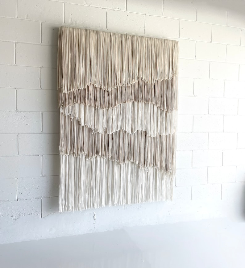Op de afbeelding: Een grote wandhanger gemaakt van geweven beige en wit garen met franjes. De hanger is gemaakt van meerdere lagen garen, waardoor een textuur en gelaagdheid ontstaat.