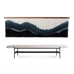 Könnte beinhalten: Ein großer Wandbehang mit dunkler Holzoberseite und einem fließenden, geschichteten Design in Schwarz-, Türkis- und Weißtönen. Darunter eine lange Bank mit hell gepolsterter Oberseite und schwarzen Metallbeinen.