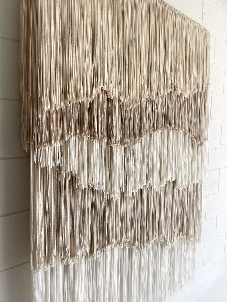 Op de afbeelding: Een beige en witte macram&eacute; wandhanger met een gelaagd franje ontwerp. De franjes zijn gemaakt van garen en zijn gerangschikt in een cascade patroon.
