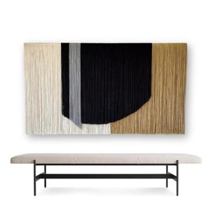 Könnte beinhalten: Ein gewebter Wandbehang in Beige, Grau, Schwarz und Braun mit einer geschwungenen Form in der Mitte. Der Wandbehang ist aus Garn gefertigt und hängt an einer weißen Wand über einer beigefarbenen Bank mit schwarzen Beinen.