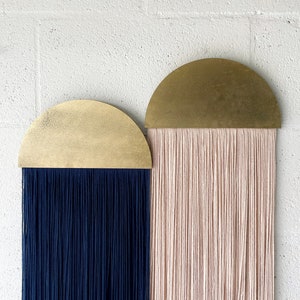 Könnte beinhalten: Zwei Wandbehänge mit einem halbrunden Messing-Oberteil und langen Garnsträngen. Das Garn links ist dunkelblau und das Garn rechts ist hellrosa.