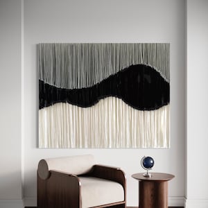 Könnte beinhalten: Ein abstrakter Wandbehang in Schwarz und Weiß mit einem wellenförmigen Design. Der Behang besteht aus Garn und wird in einem modernen Wohnzimmer mit einem Holzstuhl und einem Beistelltisch ausgestellt.