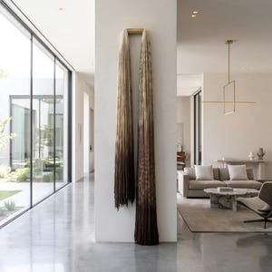 May include: A long, vertical wall hanging featuring a gradient of colors transitioning from a light cream at the top to a deep, dark brown at the bottom. The piece is composed of numerous thin, hanging strands suspended from a gold-toned metal bar.