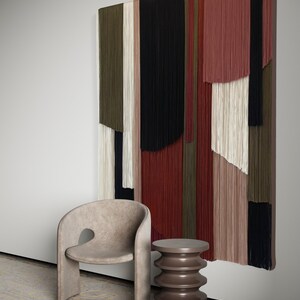 May include: A beige armchair with a unique cutout design sits next to a brown, cylindrical side table with a textured surface. A large wall hanging with colorful, vertical stripes of yarn hangs on the wall behind the furniture.