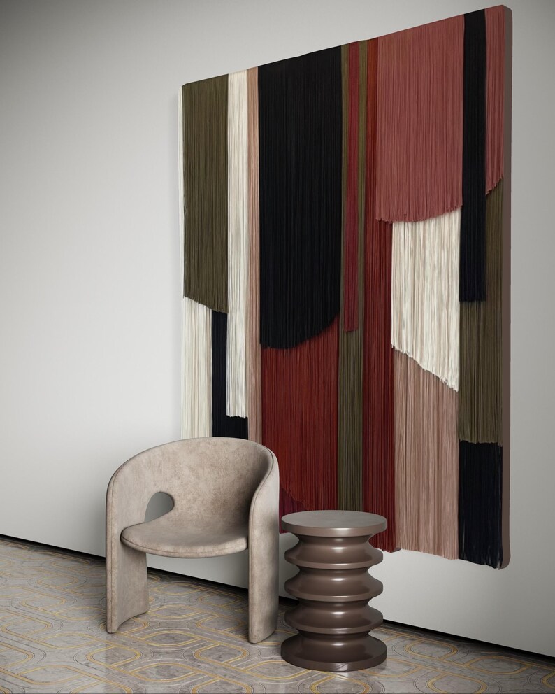 May include: A beige armchair with a unique cutout design sits next to a brown, cylindrical side table with a textured surface. A large wall hanging with colorful, vertical stripes of yarn hangs on the wall behind the furniture.