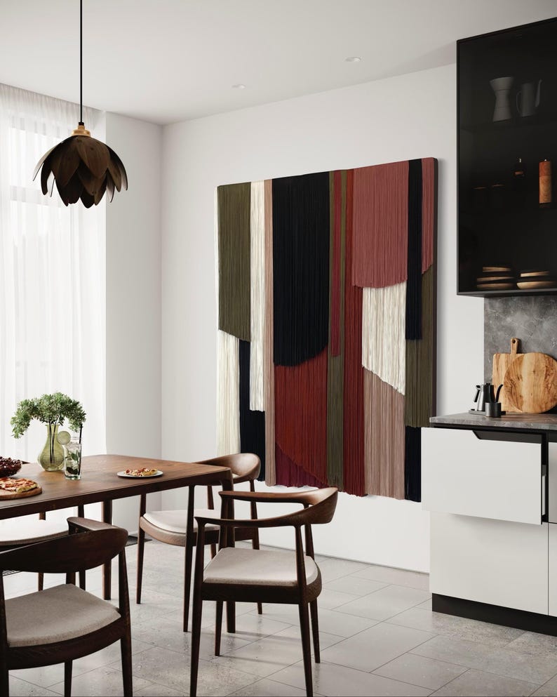 May include: A modern dining room with a wooden table and chairs. The wall features a large abstract wall hanging made of yarn in shades of red, green, black, and white.