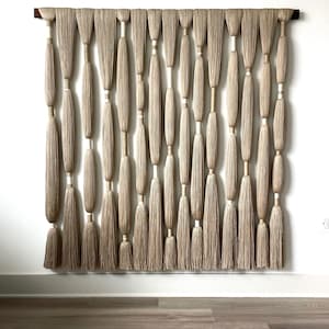 Könnte beinhalten: Ein beige-weißer Makramee-Wandbehang mit einer Holzleiste oben. Der Wandbehang besteht aus mehreren Garnsträngen, die miteinander verknotet sind, um eine Reihe von Quasten zu bilden.