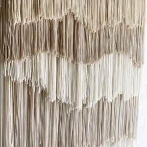 Op de afbeelding: Een wandkleed gemaakt van geweven garen met een gradi&euml;nt van beige en witte kleuren. Het garen is gerangschikt in een reeks van cascade-lagen, waardoor een textuur en een visueel aantrekkelijk ontwerp ontstaat.