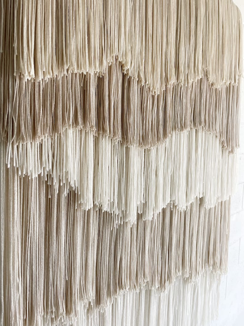 Op de afbeelding: Een wandkleed gemaakt van geweven garen met een gradi&euml;nt van beige en witte kleuren. Het garen is gerangschikt in een reeks van cascade-lagen, waardoor een textuur en een visueel aantrekkelijk ontwerp ontstaat.