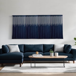 May include: A navy blue sectional sofa with white pillows and a matching wall hanging. The wall hanging features a series of navy blue tassels. A wooden coffee table and a small plant are also visible.