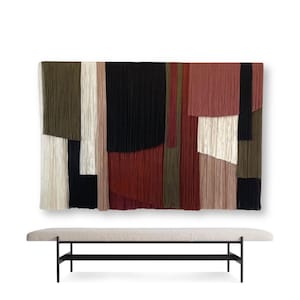 May include: A large abstract wall hanging made of yarn in shades of red, green, black, and white. The hanging is displayed above a long, rectangular bench with a light gray fabric cushion and black metal legs.