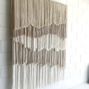 Op de afbeelding: Een wandkleed gemaakt van beige en wit garen met een gelaagd, cascade-achtig franjemotief.