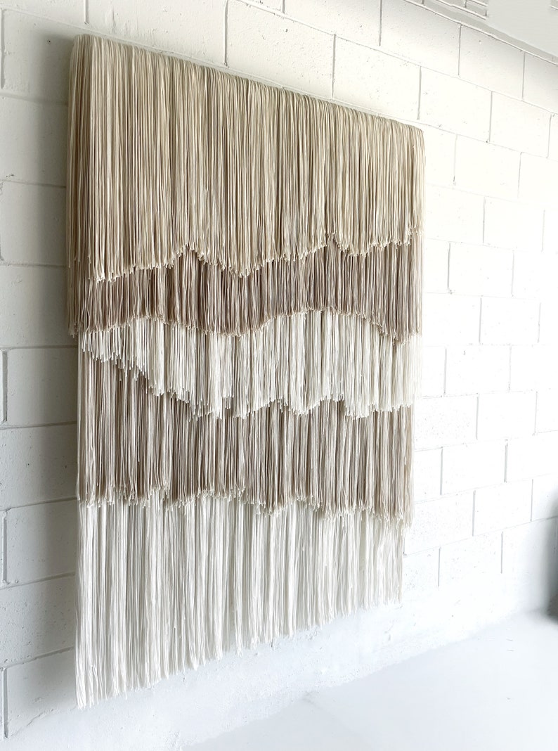 Op de afbeelding: Een wandkleed gemaakt van beige en wit garen met een gelaagd, cascade-achtig franjemotief.