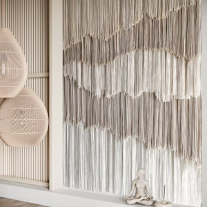 Op de afbeelding: Een wit keramiek beeldje van een persoon die in meditatieve houding zit, met een bruine wierookbrander en een wierookstokje ervoor. Het beeldje staat voor een muur met een beige en witte macram&eacute; wanddecoratie.