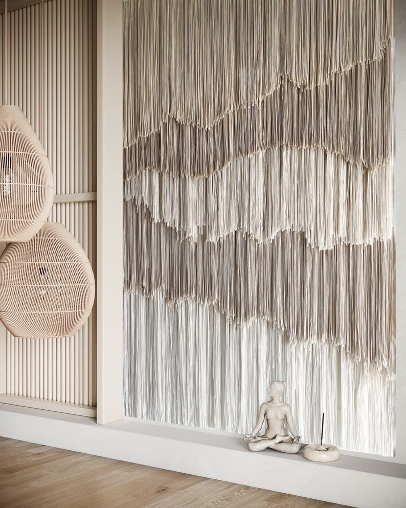 Op de afbeelding: Een wit keramiek beeldje van een persoon die in meditatieve houding zit, met een bruine wierookbrander en een wierookstokje ervoor. Het beeldje staat voor een muur met een beige en witte macram&eacute; wanddecoratie.
