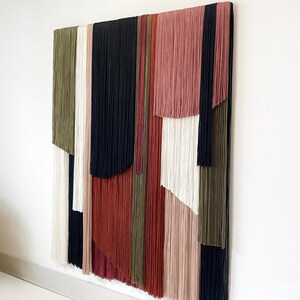 May include: A wall hanging made of yarn in various colors, including black, white, red, pink, and green. The yarn is arranged in vertical strips, creating a geometric pattern.