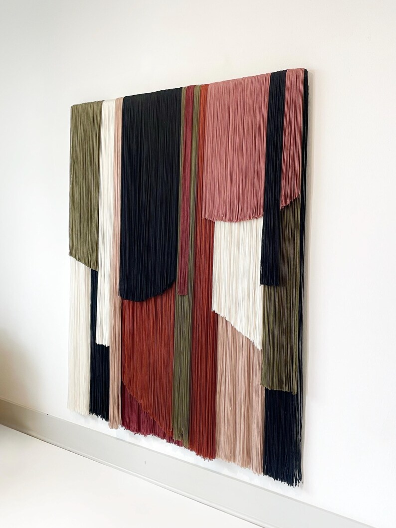 May include: A wall hanging made of yarn in various colors, including black, white, red, pink, and green. The yarn is arranged in vertical strips, creating a geometric pattern.