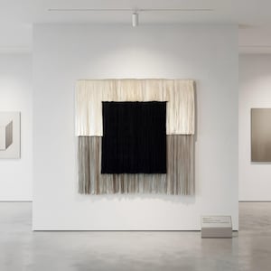 May include: A large, abstract textile artwork with a central black square, framed by white and gray fringed sections. The piece hangs on a white gallery wall, with other artworks visible in the background. The artwork is a modern, minimalist design.
