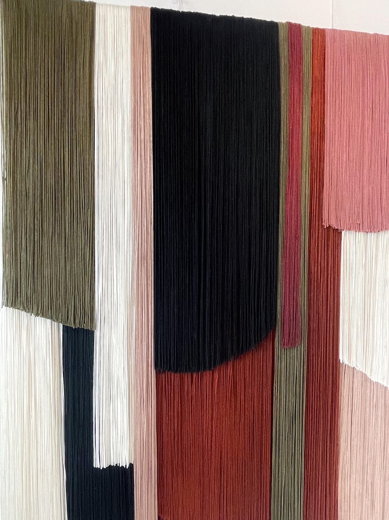 May include: A wall hanging made of yarn in various colors, including green, white, black, red, and pink. The yarn is arranged in vertical stripes, creating a geometric pattern.