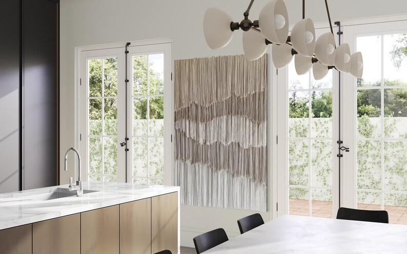 Op de afbeelding: Een moderne keuken met witte marmeren aanrechtbladen en een kraan van roestvrij staal. De keuken heeft een groot raam met uitzicht op een weelderige groene tuin. Een geweven wandtapijt voegt een vleugje bohemian stijl toe aan de ruimte.
