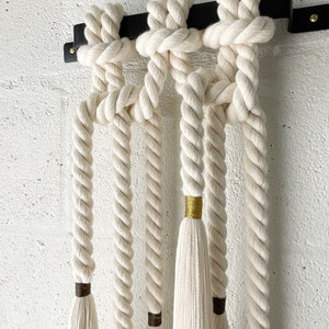 Könnte beinhalten: Ein Wandbehang aus weißem Seil mit goldenen und braunen Quasten. Das Seil ist verknotet und an einer schwarzen Metallstange befestigt.